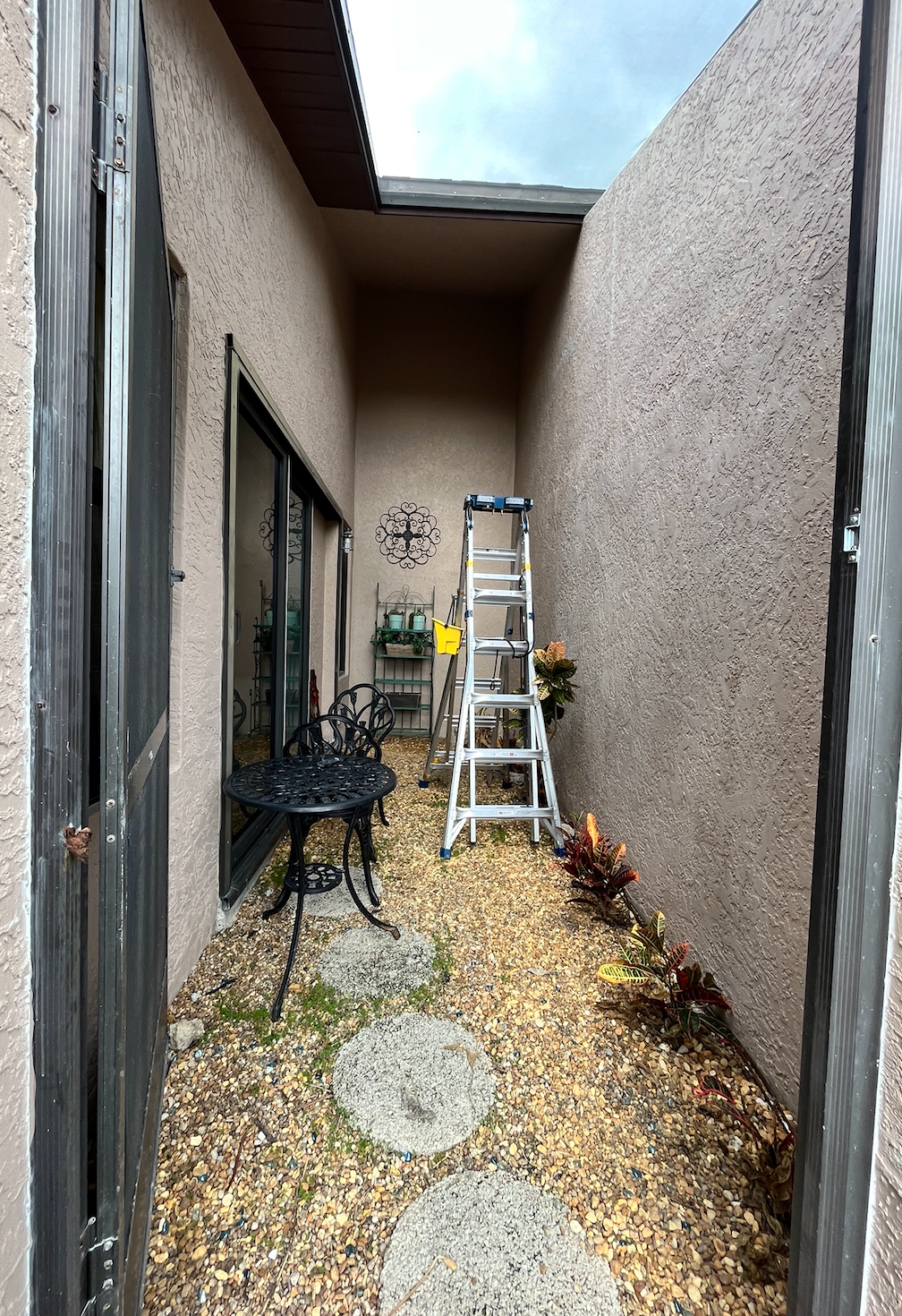 Courtyard with a black table and chair, gravel ground, and ladder against a wall.