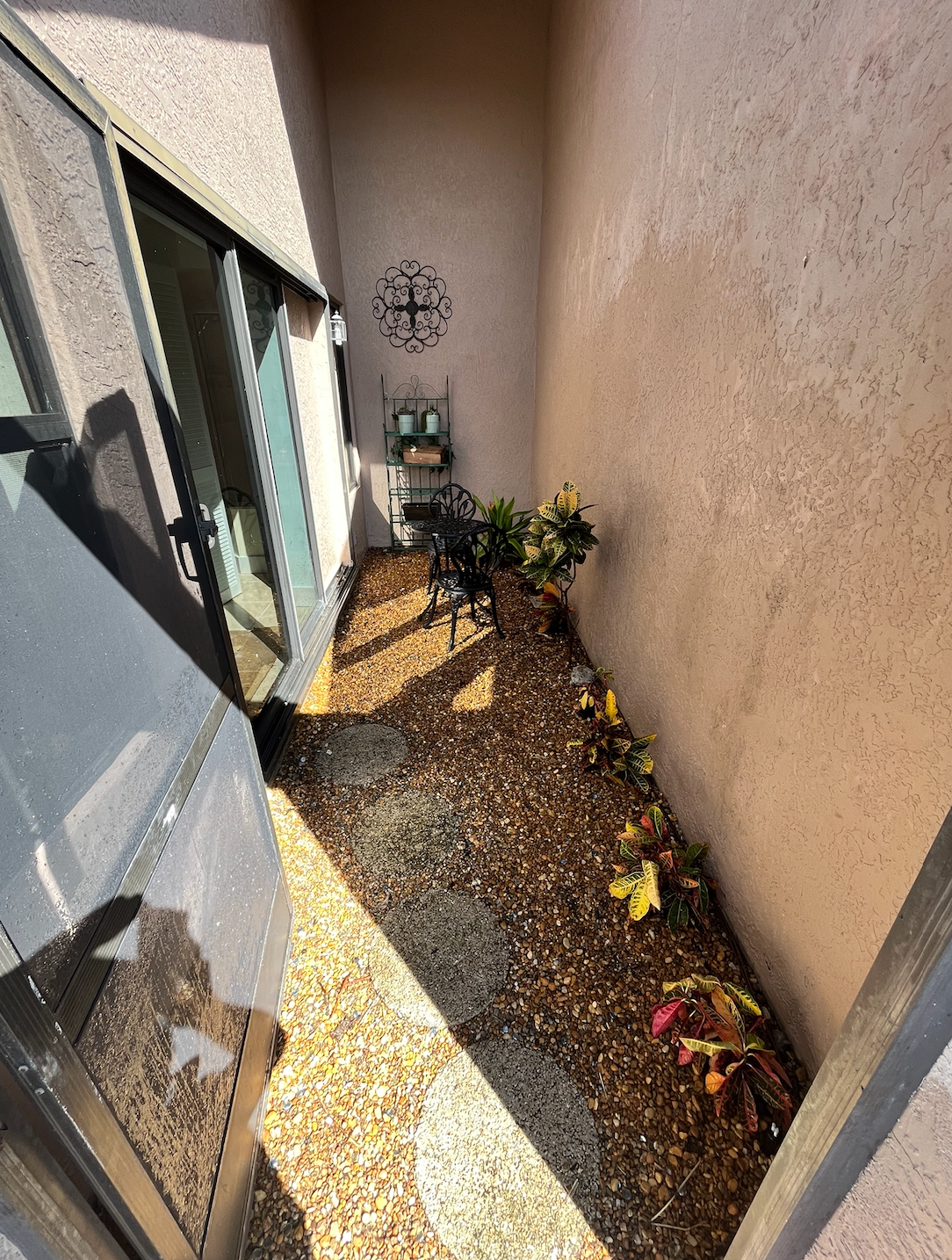 Narrow outdoor space with stone path, plants, decorative wall cross, and sliding glass door.