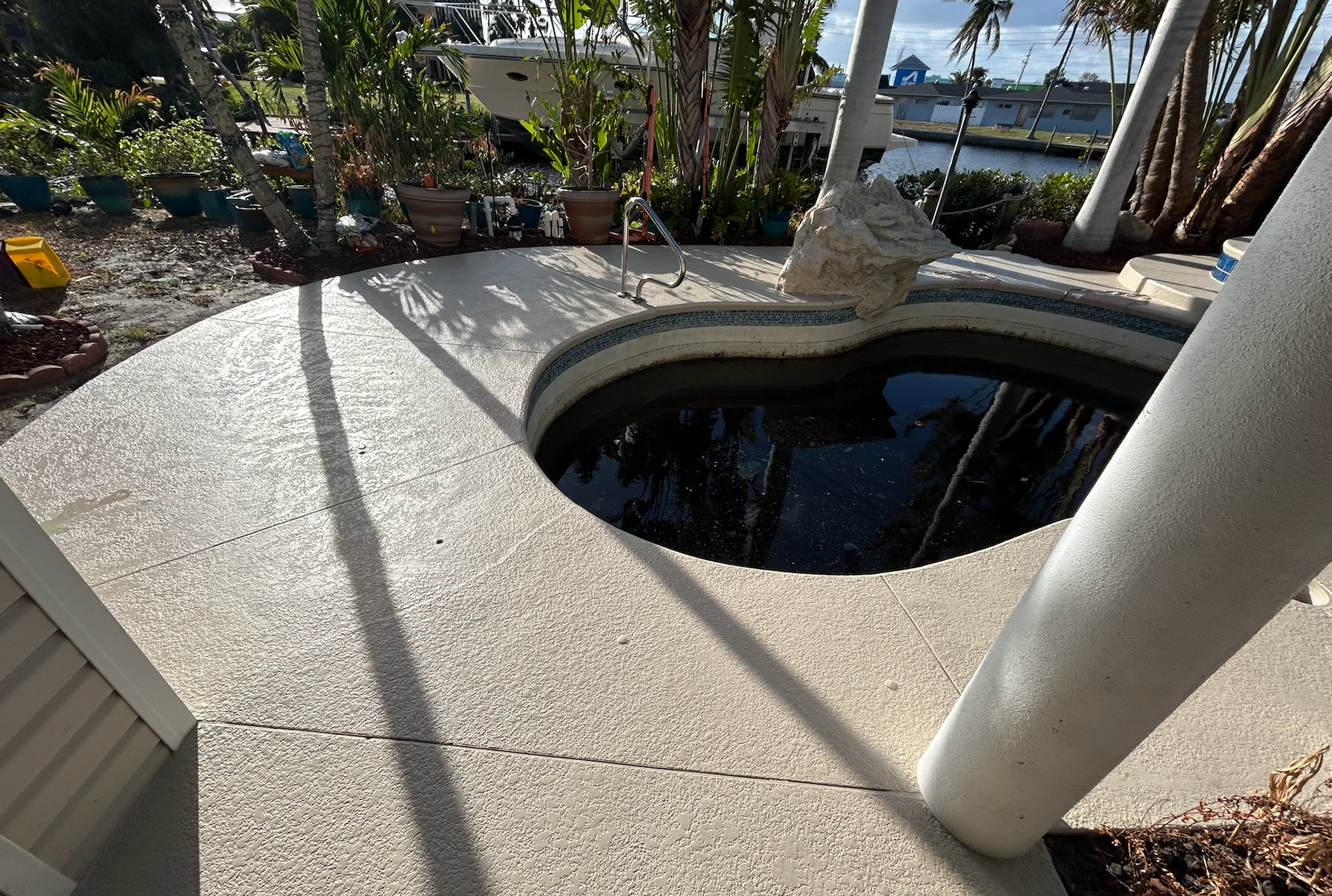 Concrete patio surrounds a dark-colored, heart-shaped pool. Trees and a boat are visible in the background.