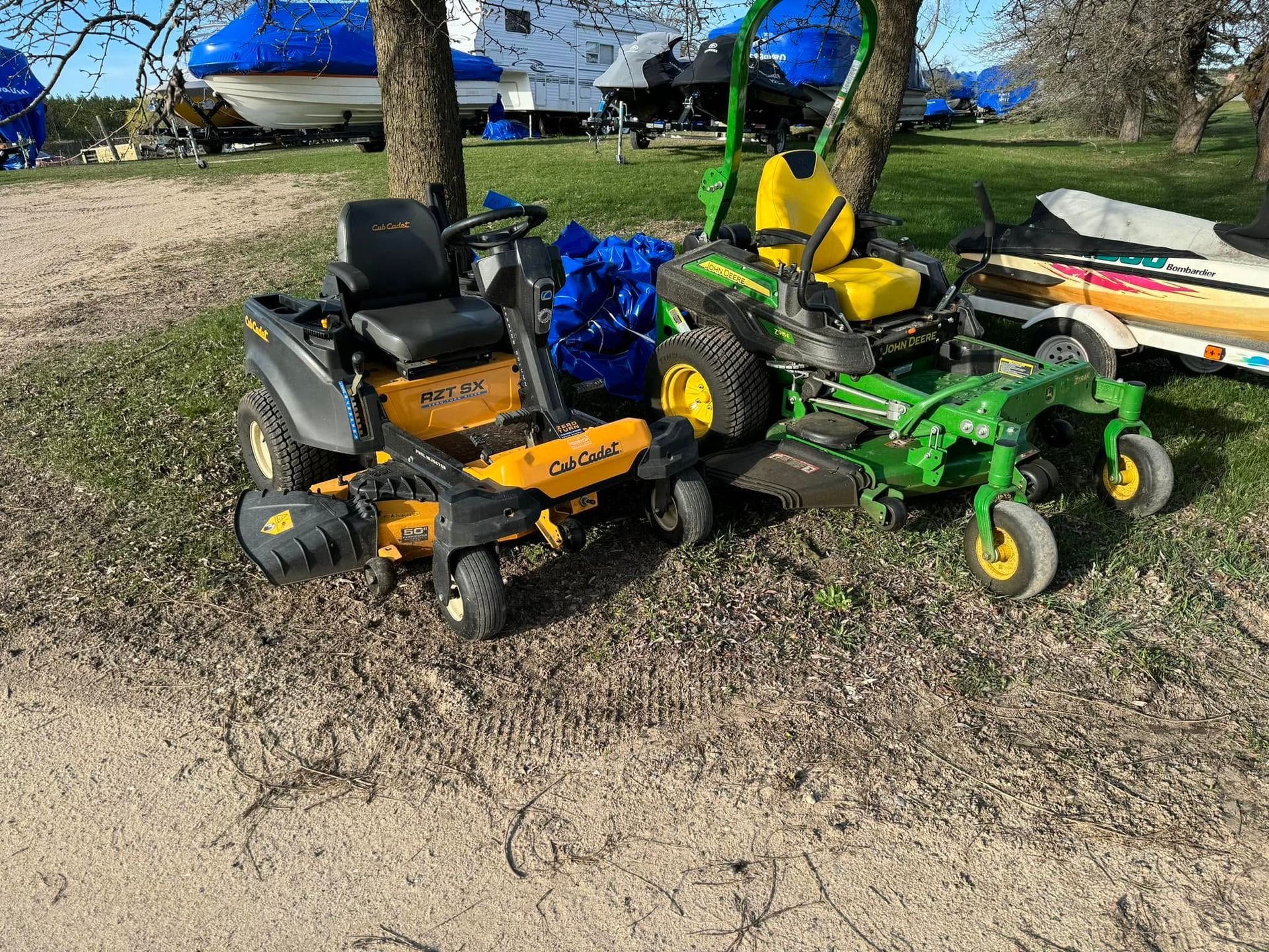 Two riding lawn mowers, one orange and one green, parked on grass near a farmyard.