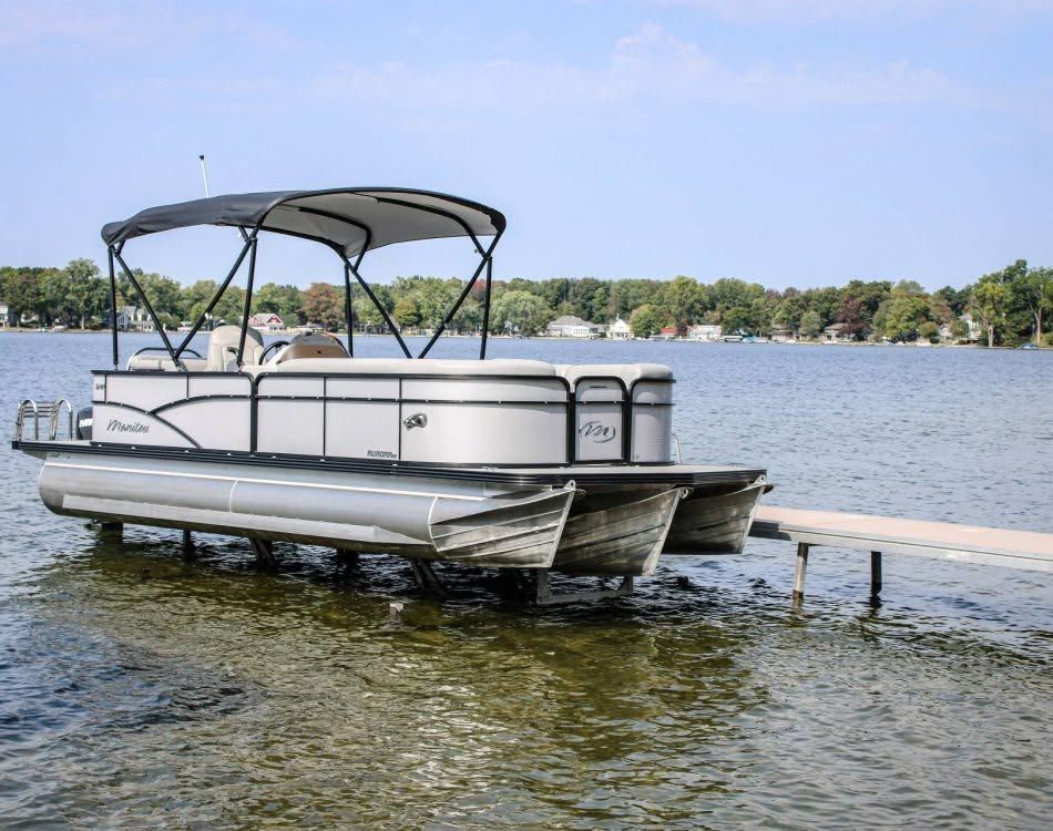Pontoon boat moored by a dock on a calm lake under a canopy, with trees on the shore behind it