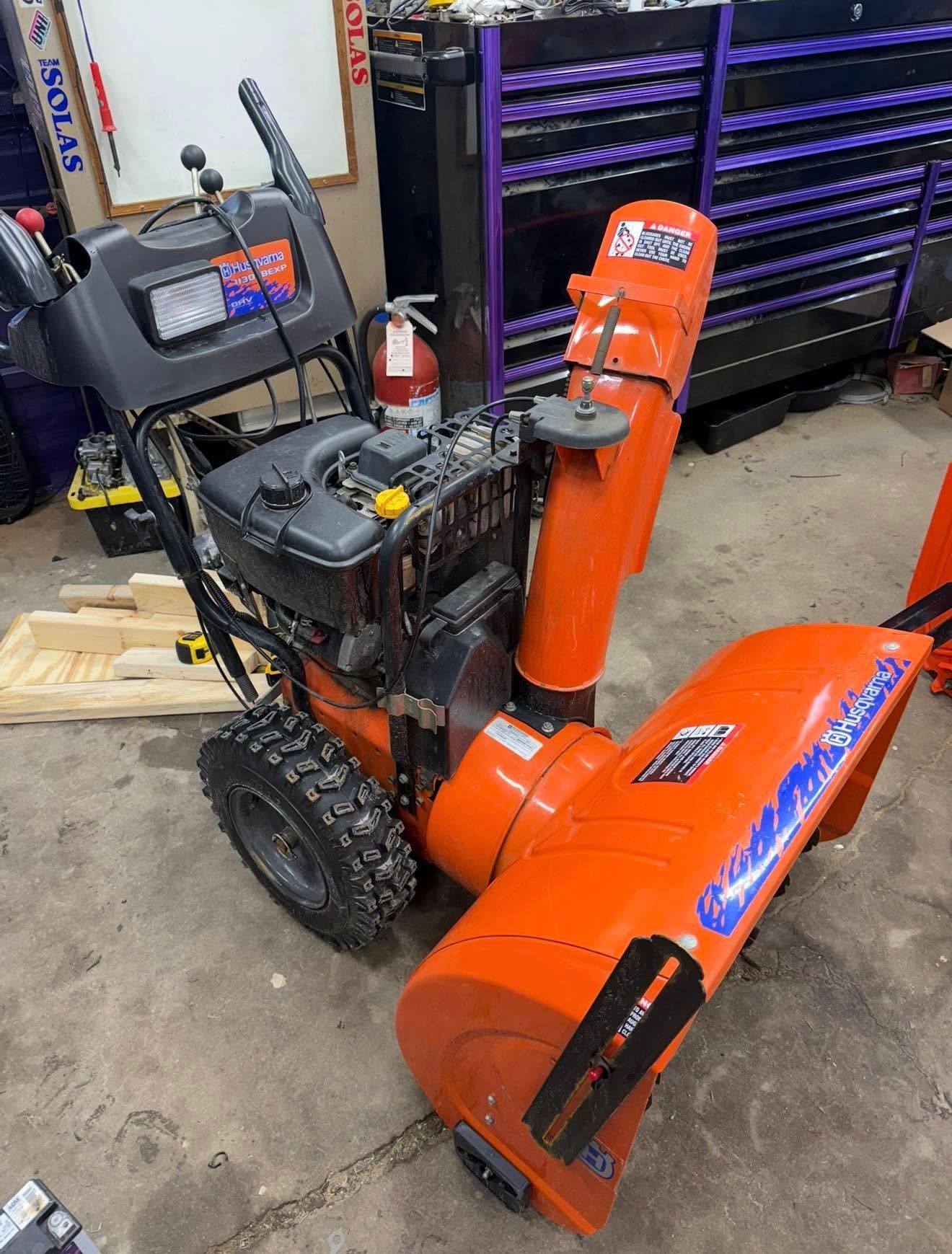 Orange snow blower in a workshop, with a black engine and wheels beside a tool cabinet.