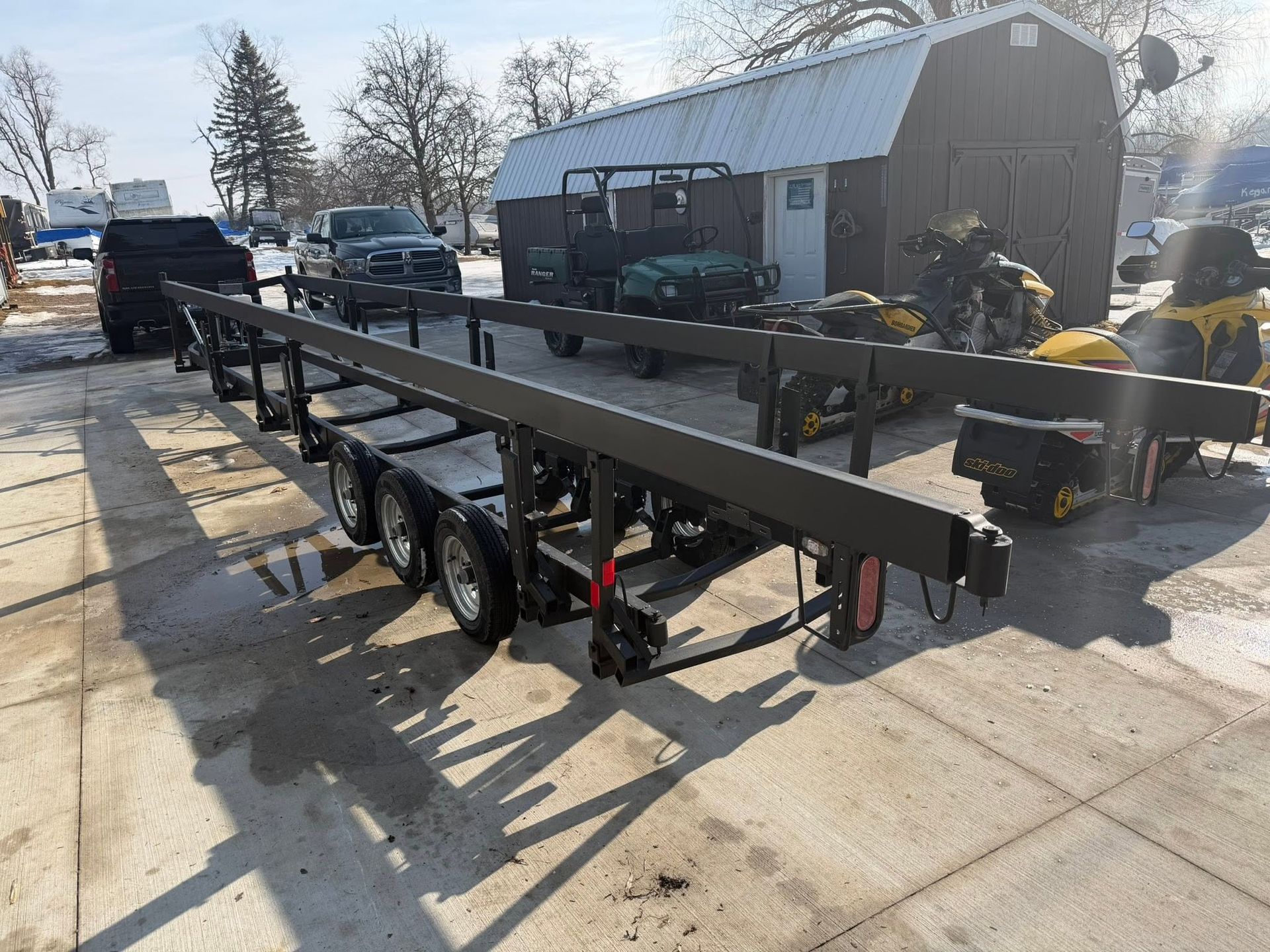 Black flatbed trailer in a driveway beside a workshop, with trucks and equipment in the background.