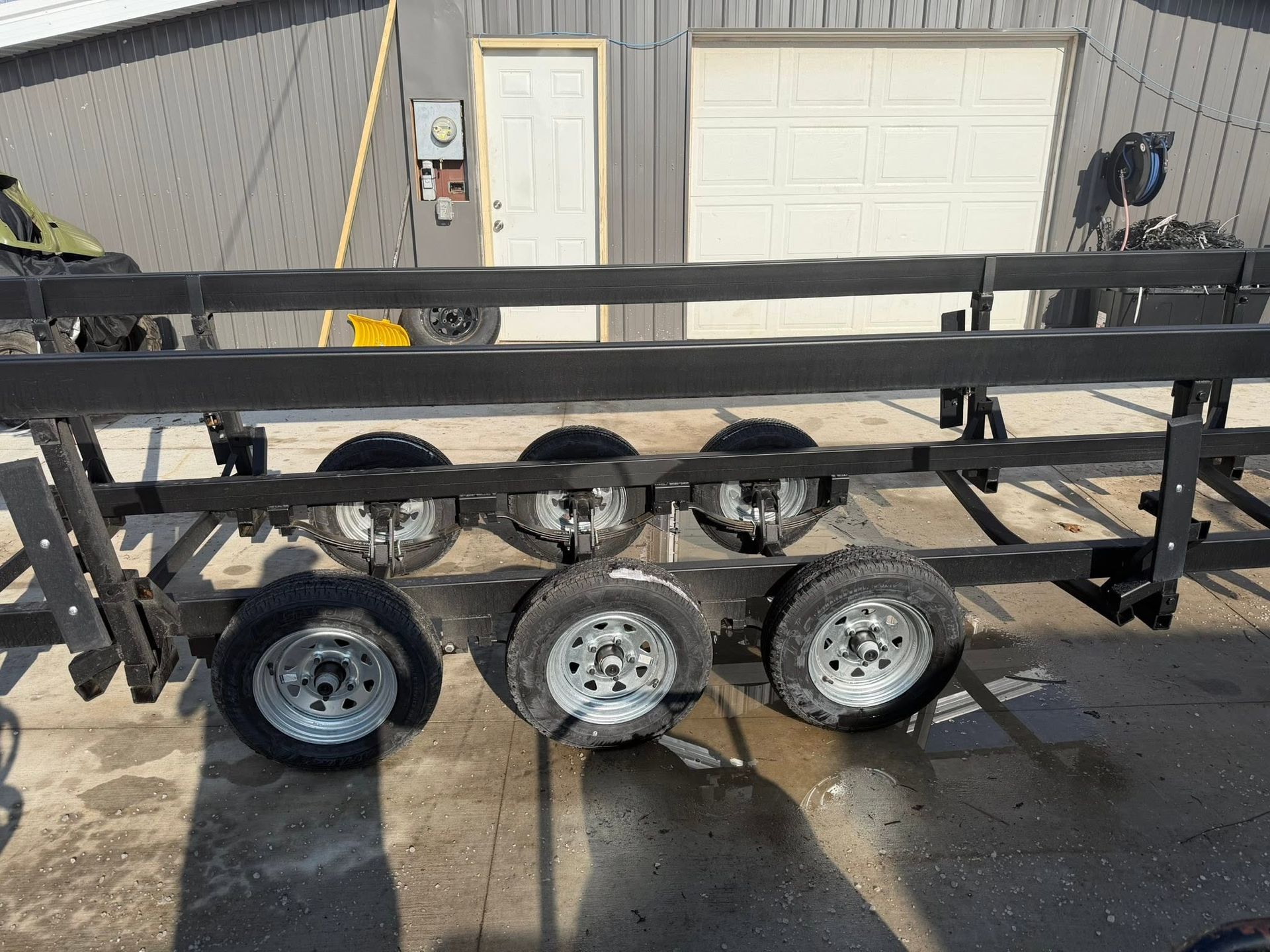 Black metal trailer frame with five wheels parked outside a garage door
