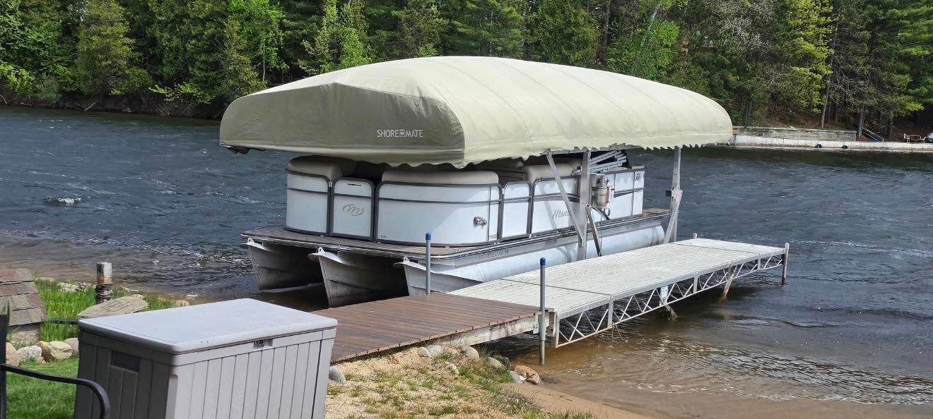 Pontoon boat with a tan canopy docked by a rocky river with forested shoreline