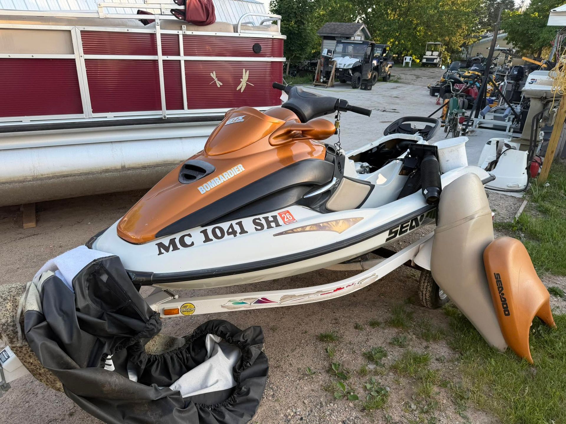 Orange and white jet ski with visible damage, parked on a dirt lot near a red truck and trees.