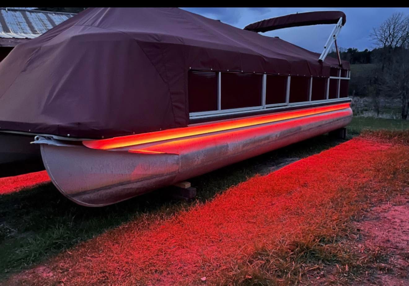 Covered pontoon boat with red LED lights glowing on grass at dusk