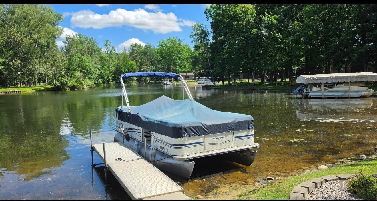 Covered pontoon boat docked at a lake beside a wooden pier, with trees and reflections on the water.