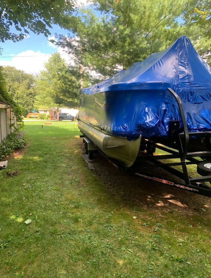 Boat covered with a blue tarp on a trailer in a grassy yard, beside trees and a fence.