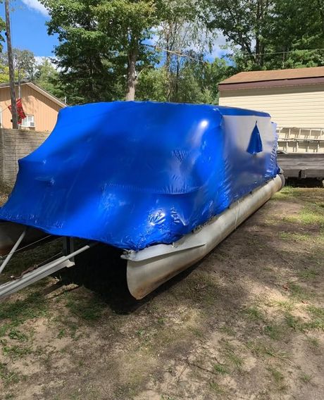 Blue tarp-covered pontoon boat parked in a yard beside a house and trees