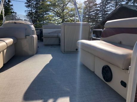 Tan pontoon boat seating on a sunny deck with lake and pine trees in the background