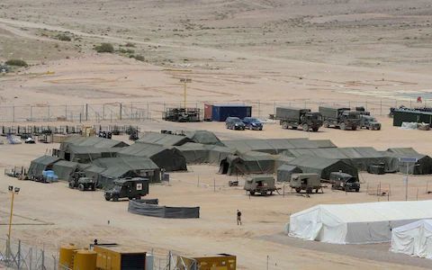 A desert military encampment with green tents, parked trucks, and a white tent in the foreground under a clear sky.