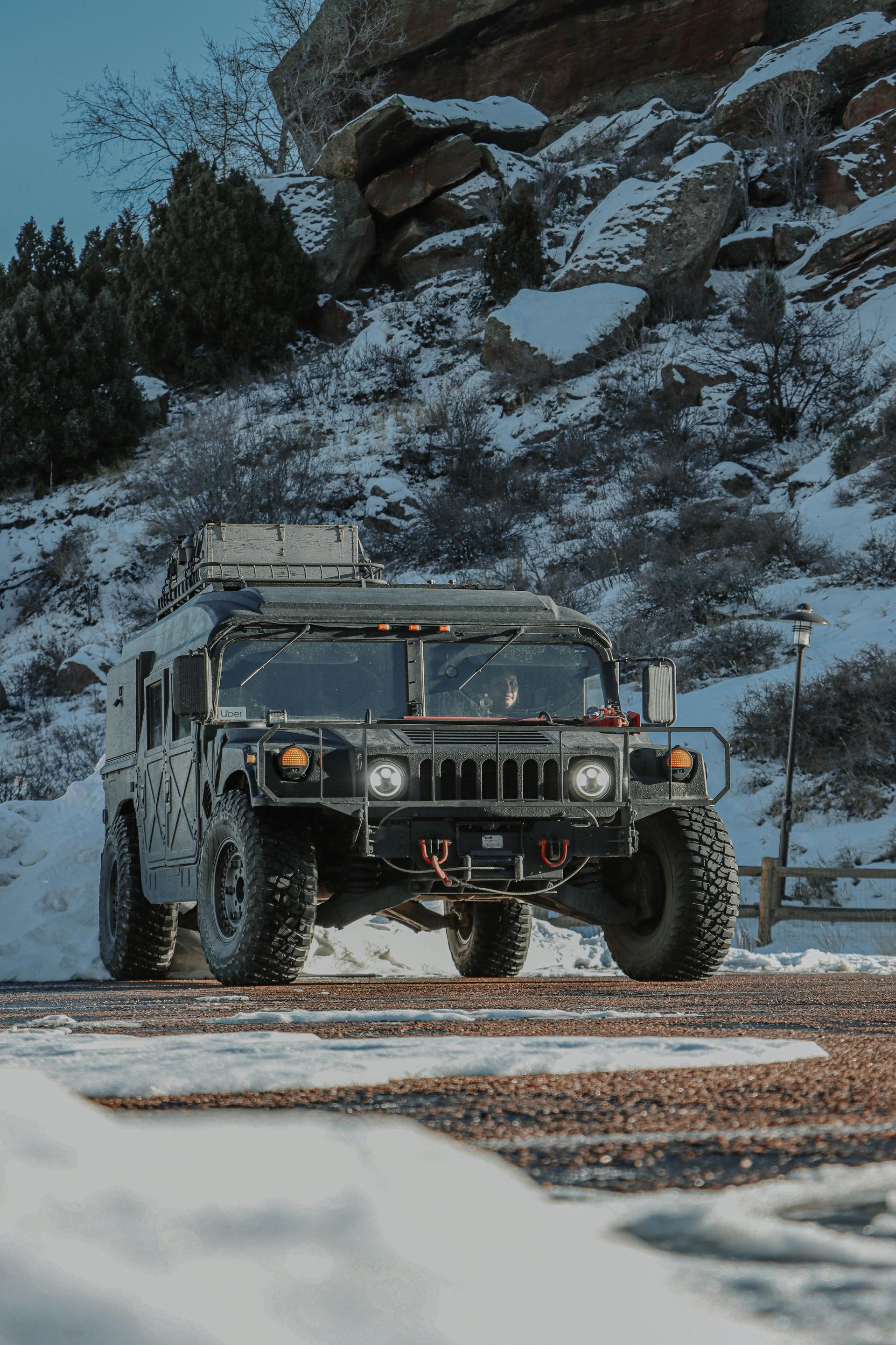 A dark-colored Humvee parked on a snowy, gravel path in front of a rocky, snow-dusted hillside.