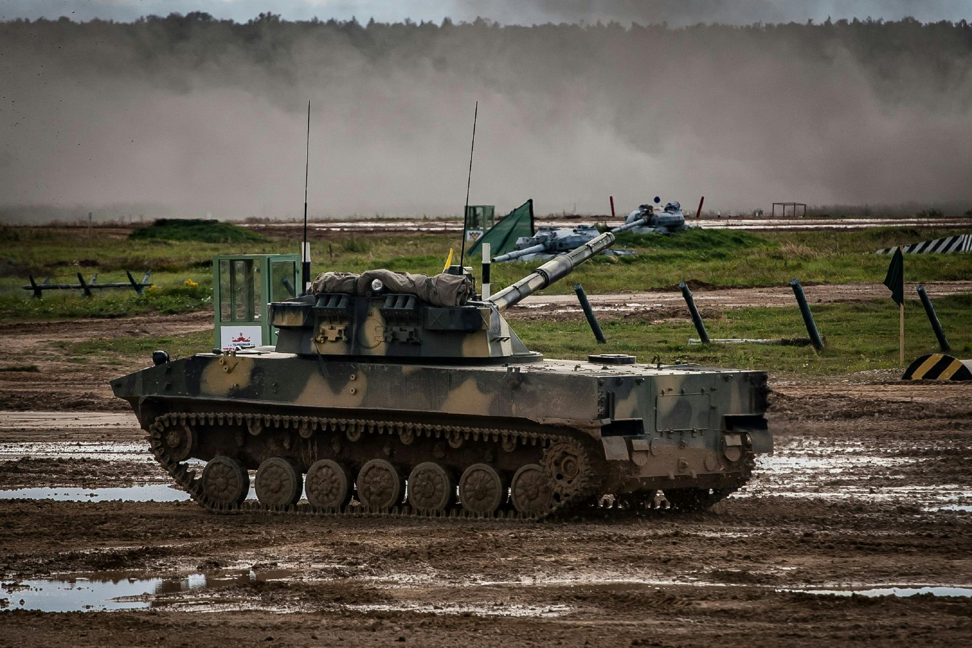 A camouflage-patterned tank maneuvers across a muddy field under a cloudy, overcast sky.
