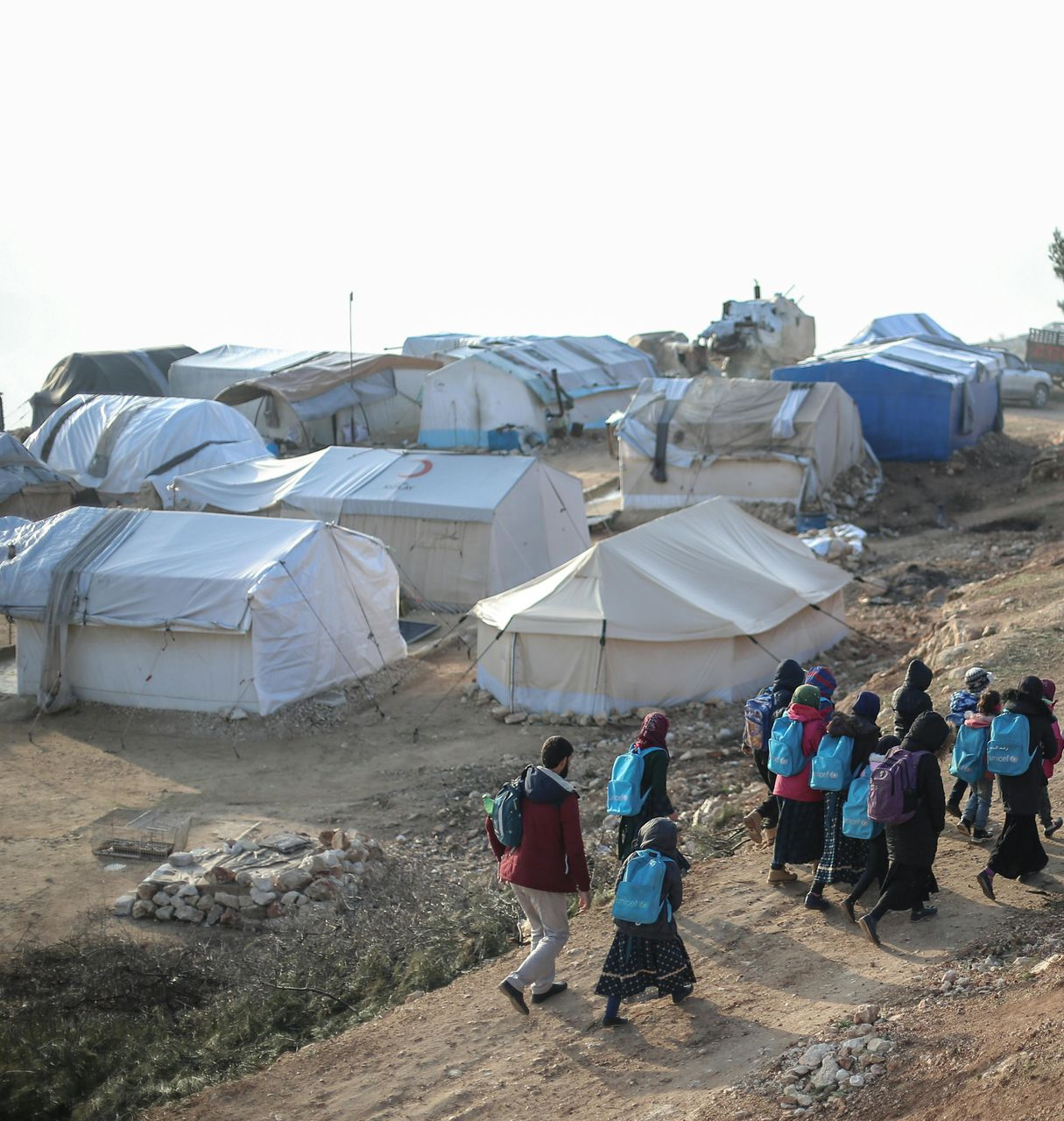 People with blue backpacks walk along a path in a settlement of makeshift white and blue tents on a rocky hillside.