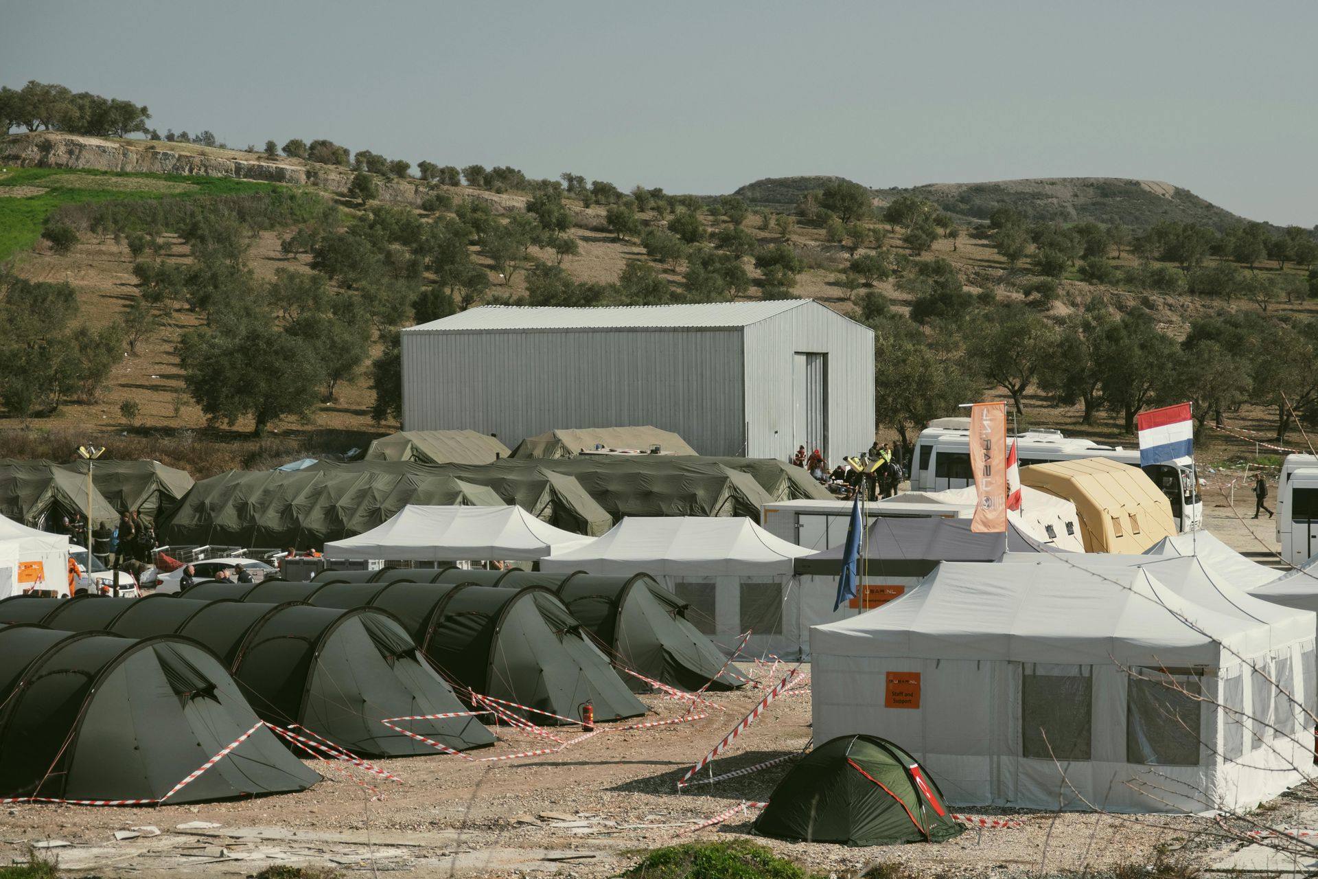 A field camp with rows of green military-style tents and white medical or administrative shelters in a hilly landscape.