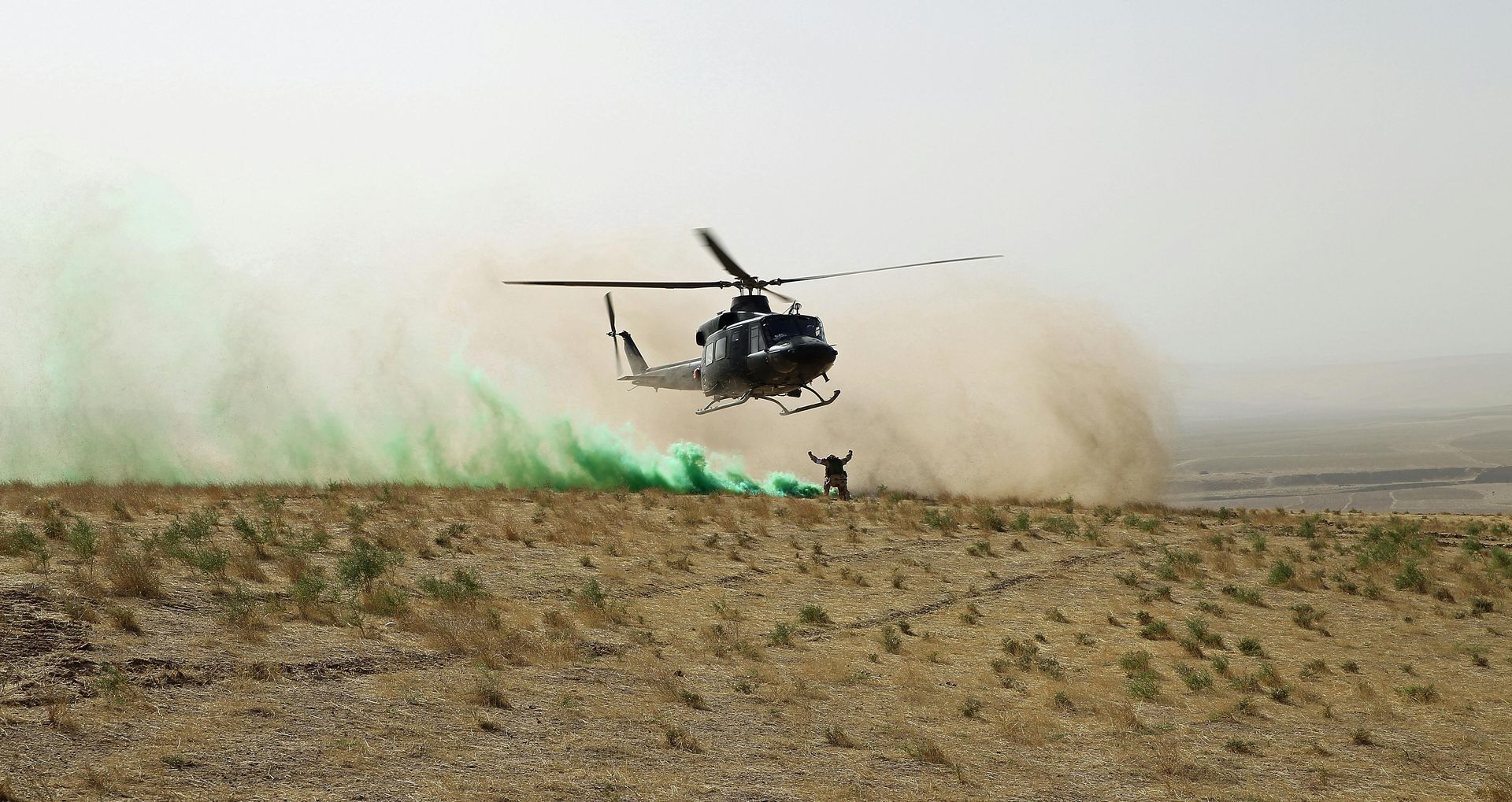 A helicopter hovers low over a dusty, arid landscape, marked by a plume of green smoke.