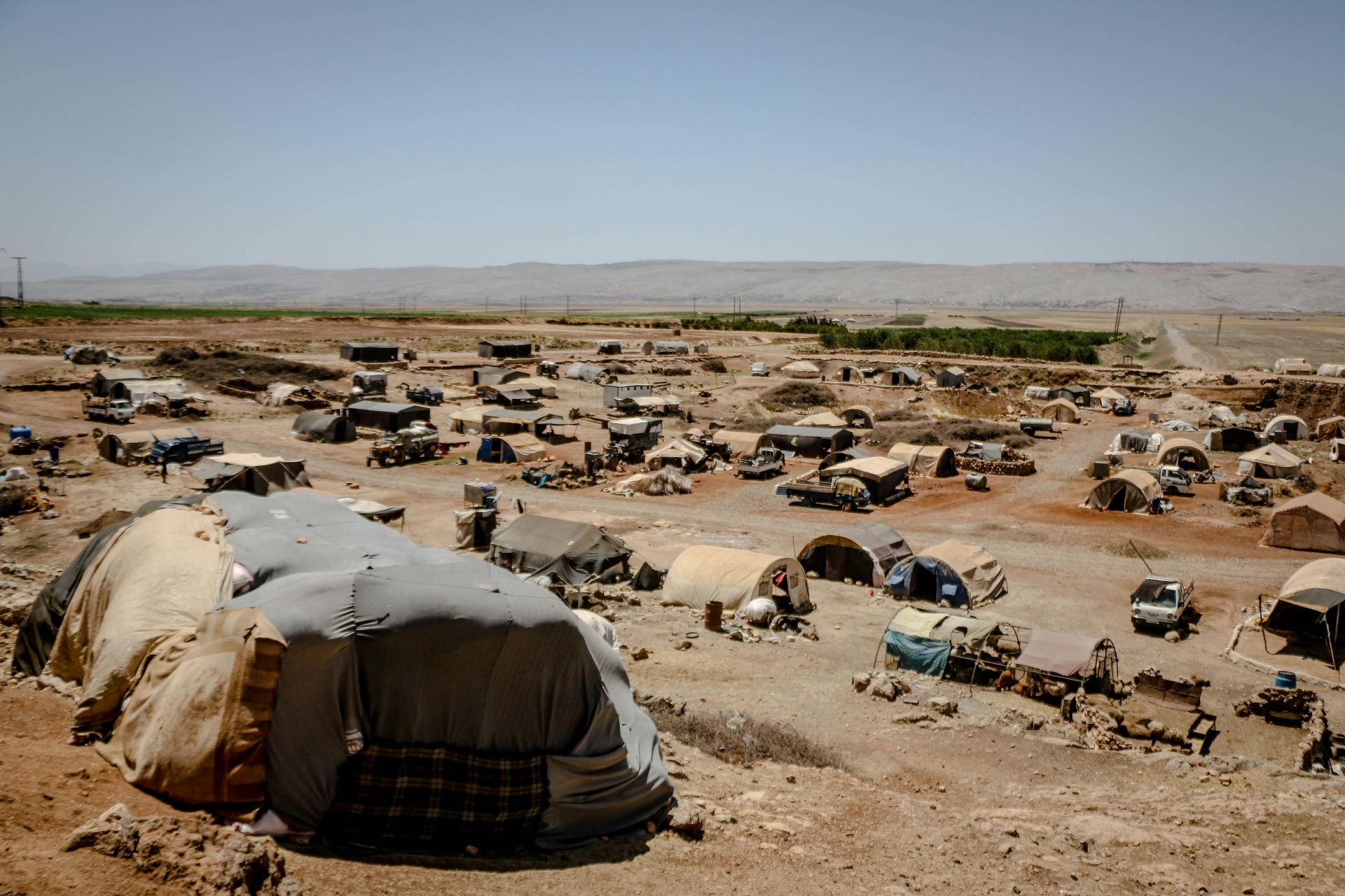 A wide, high-angle view of a sprawling refugee camp in a dry, barren landscape with many makeshift tents and shelters.