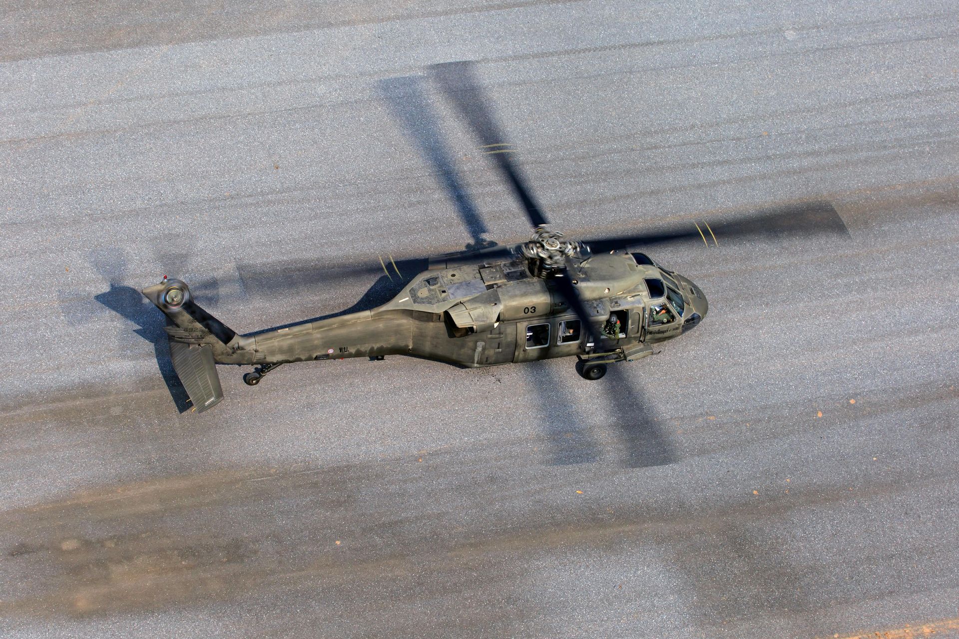 An olive-drab military helicopter flying low over a paved gray runway, seen from an overhead perspective.