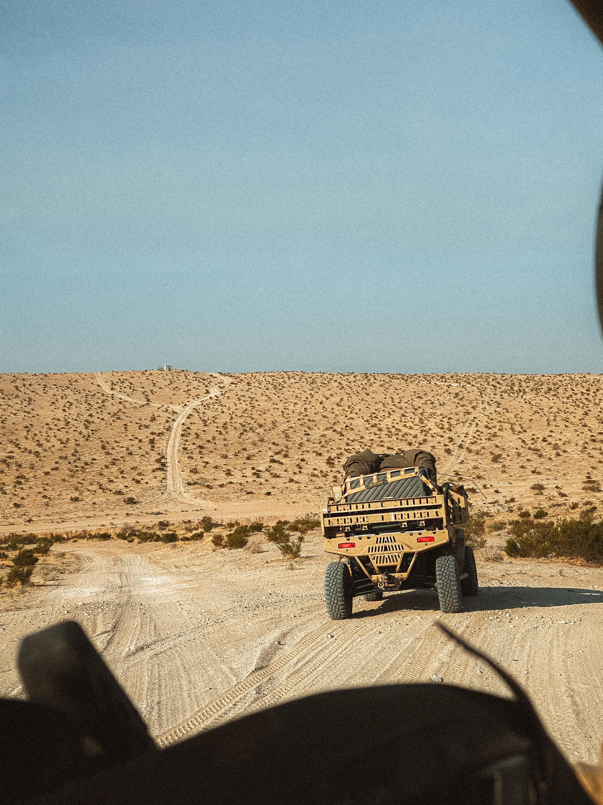 A tan military vehicle drives across a sparse, rocky desert landscape under a clear blue sky.