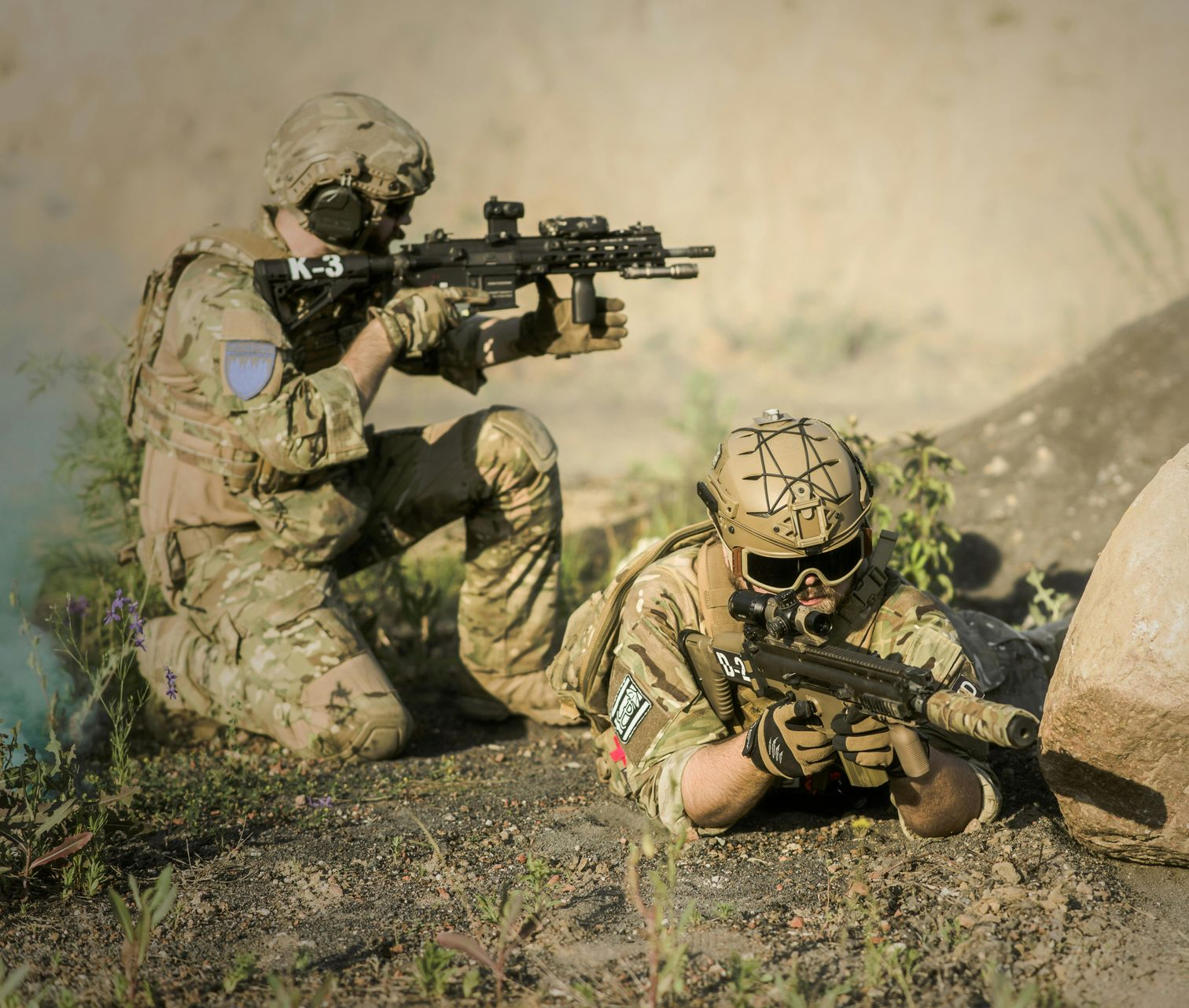 Two soldiers in camouflage gear, one kneeling and one prone, aim their rifles in a training or tactical setting.