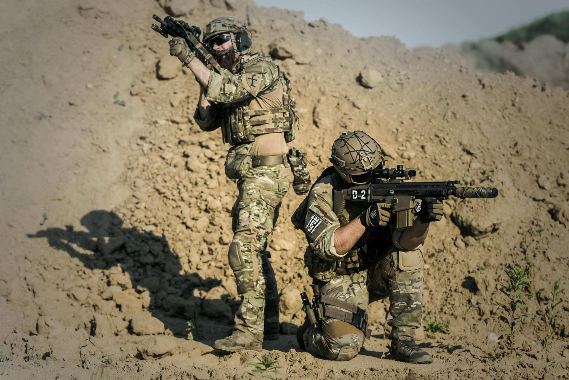 Two soldiers in camouflage gear, one standing and one kneeling, aim their rifles in front of a sandy dirt mound.
