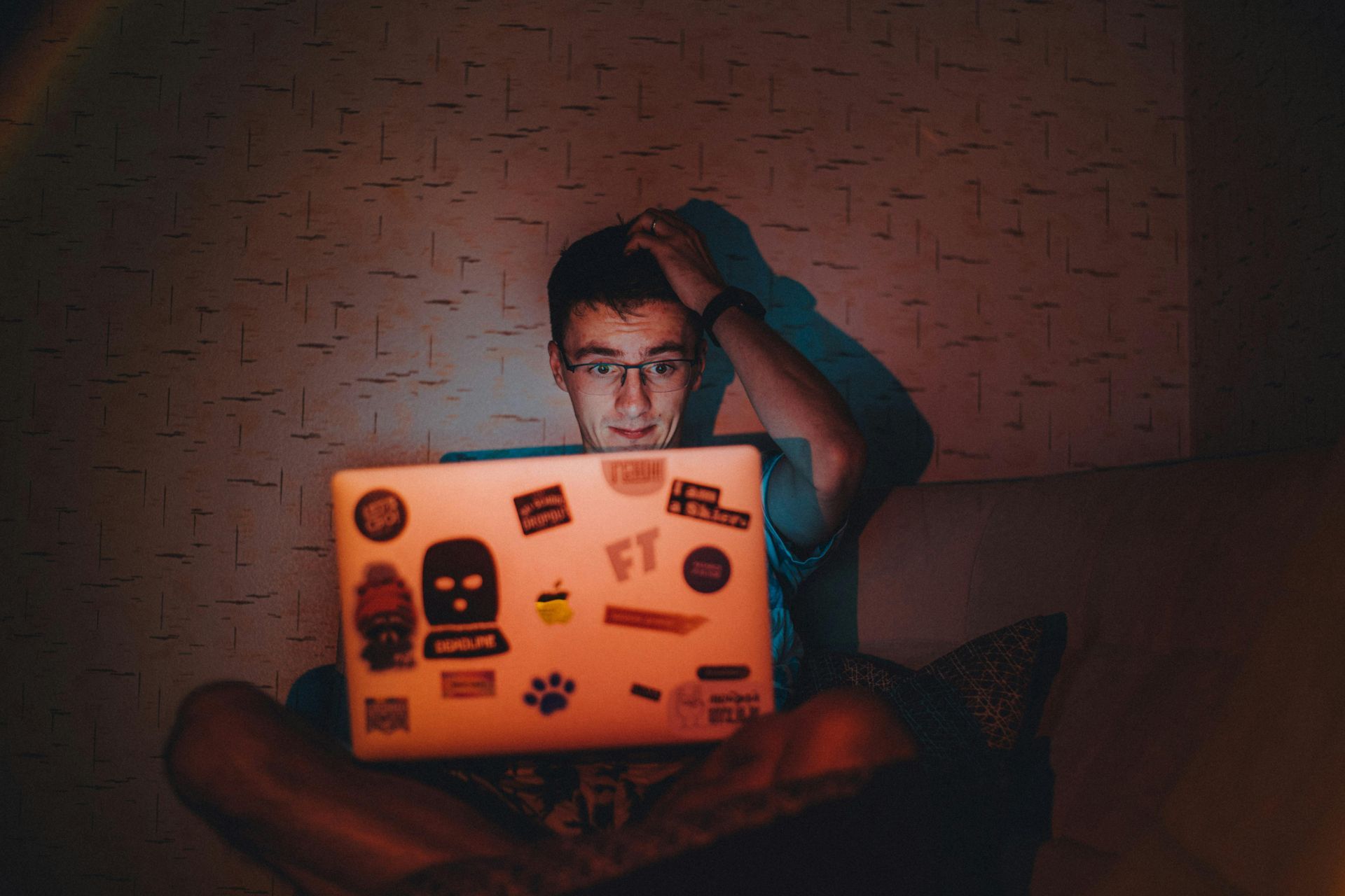 A person with glasses sits in a dim room with a sticker-covered laptop, looking frustrated while touching their hair.