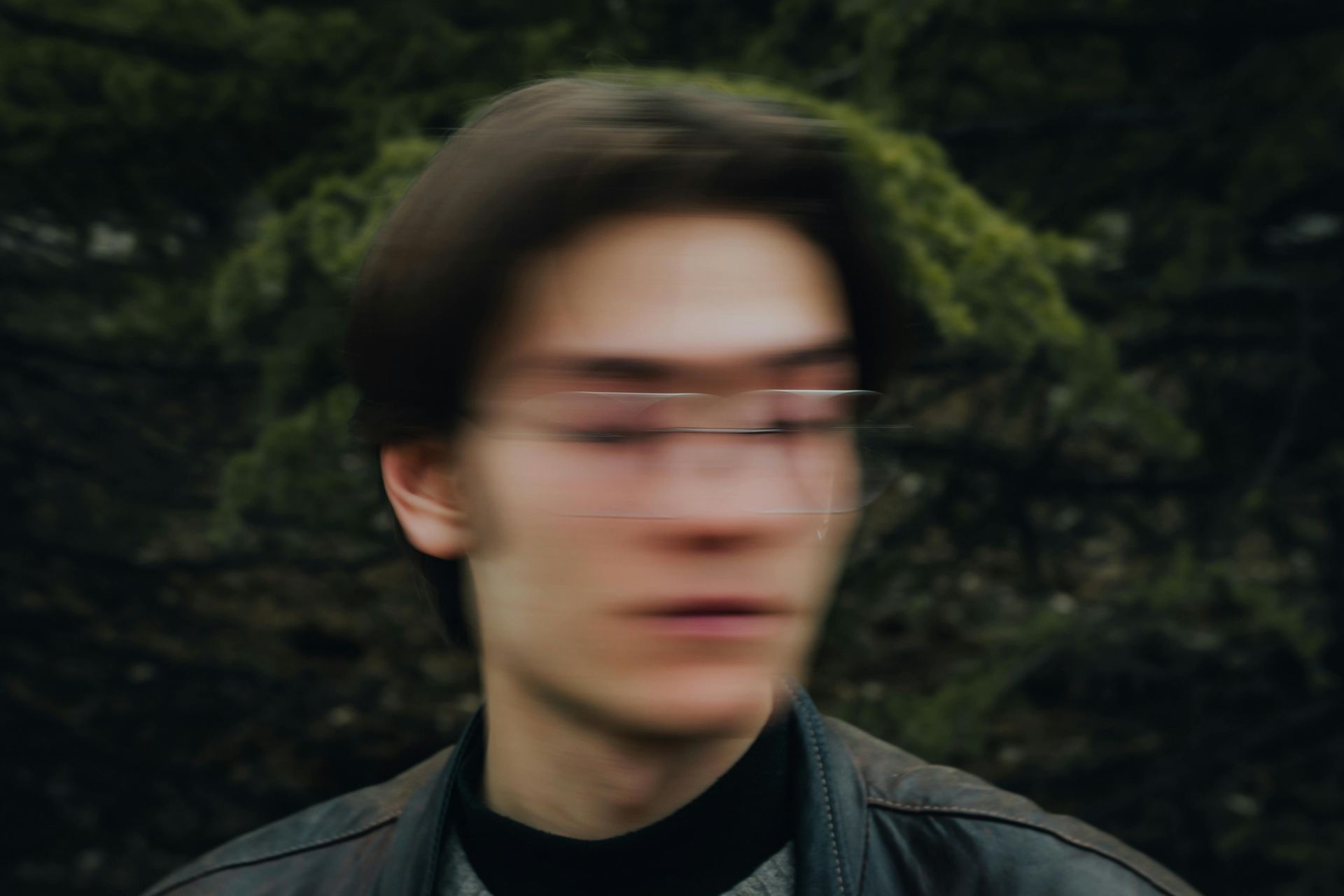 A motion-blurred, out-of-focus close-up of a person wearing glasses, centered against a dark, blurry natural background.