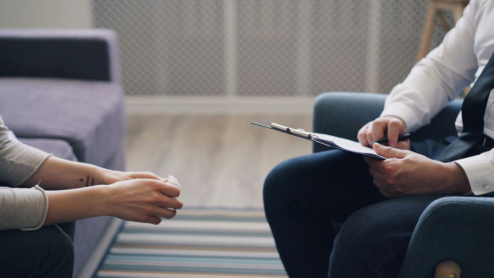 A person sits on a sofa facing a professional holding a clipboard and pen during a therapy session in a neutral office.