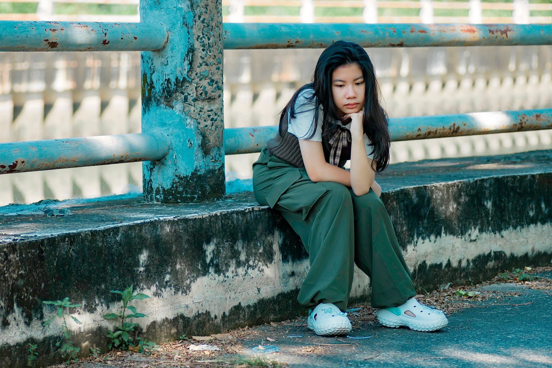 A person with long dark hair sits on a concrete ledge by a blue metal railing, looking down with a thoughtful expression.