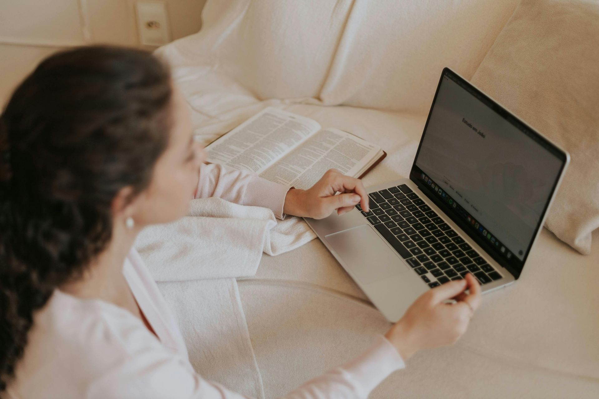 A person sits on a bed, using a laptop while an open book rests nearby.
