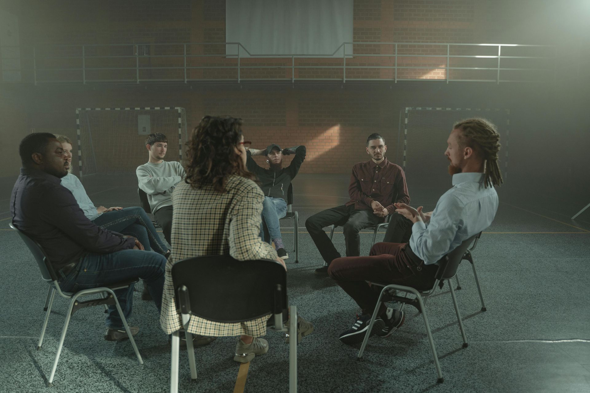 A diverse group of people sitting in a circle on chairs in a large, dimly lit, industrial-style room.