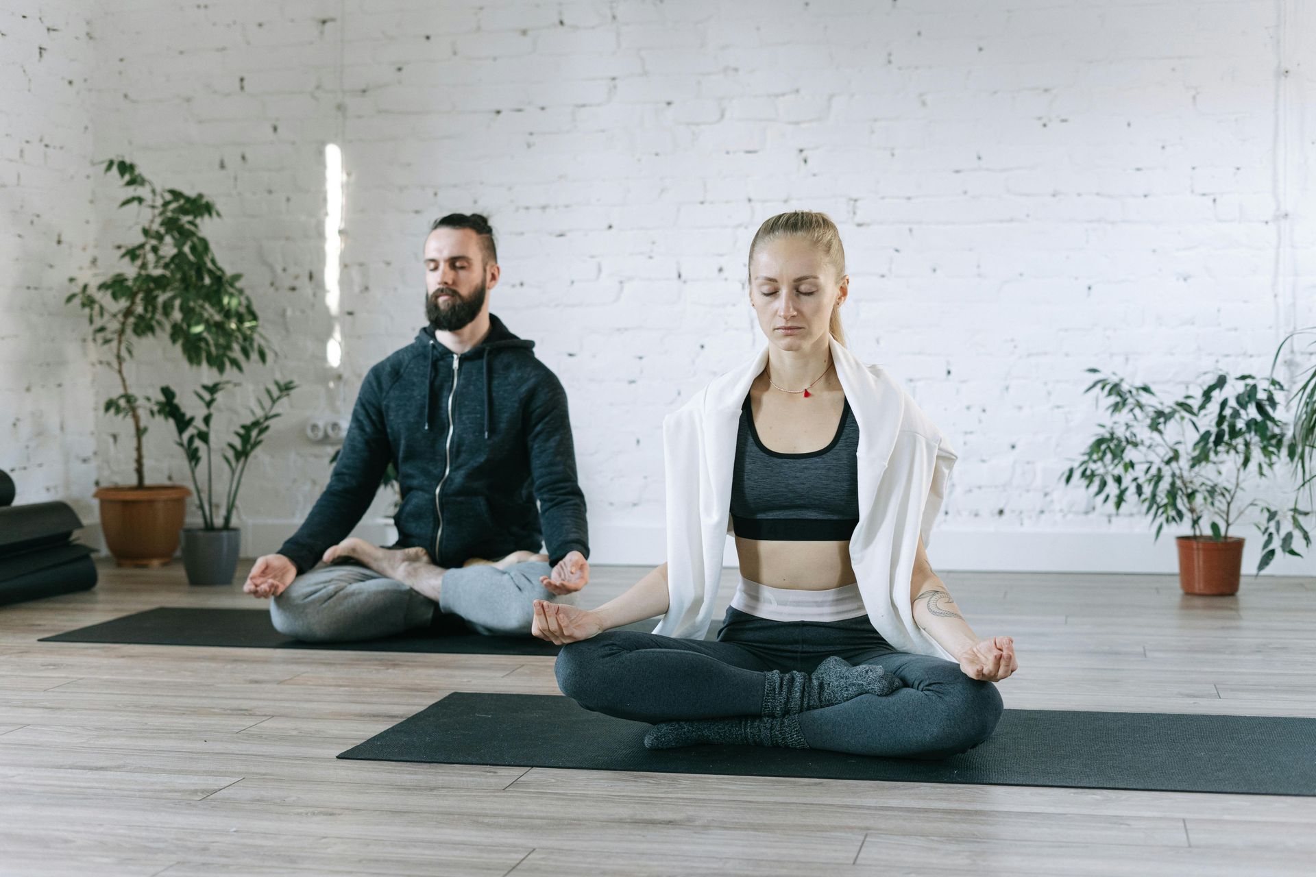 A person with a towel draped over their shoulders and a bearded person sit in a meditation pose on yoga mats.