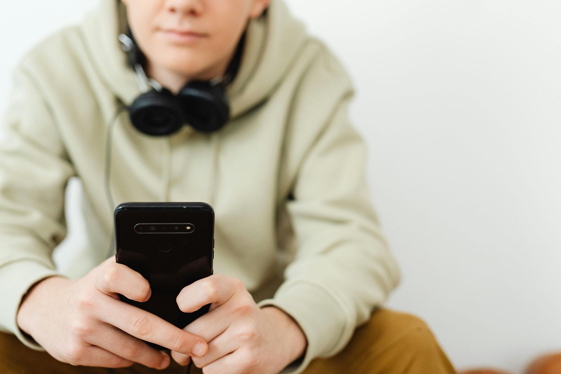A person wearing a light green hoodie and black headphones around their neck, holding a black smartphone.
