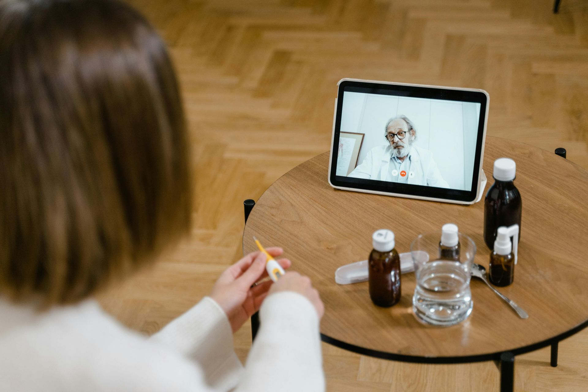 A person holds a thermometer while having a video consultation with a doctor on a tablet placed on a round table.