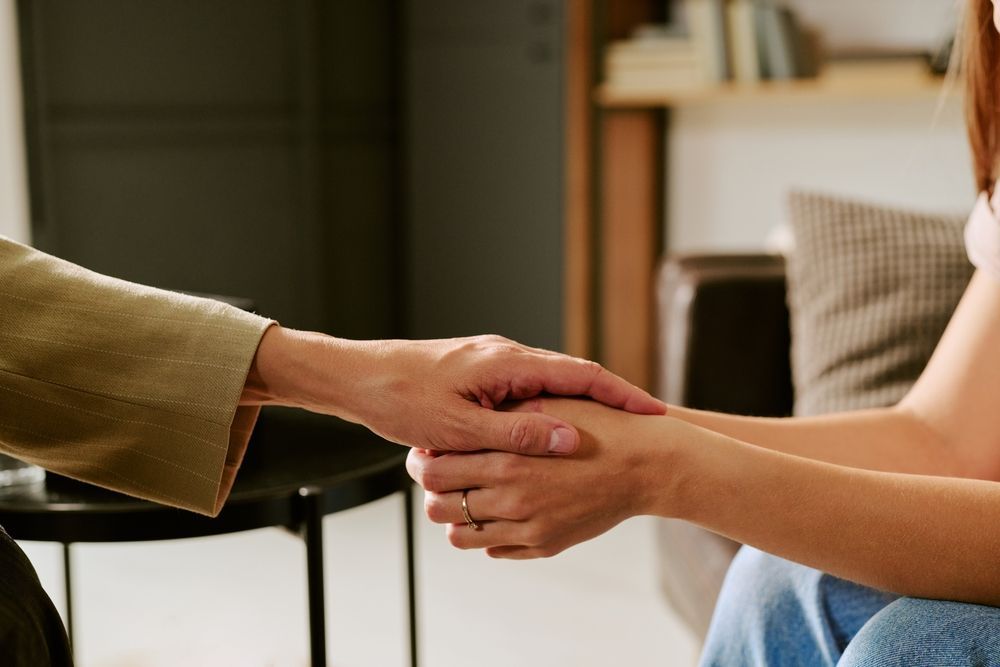 Two people hold hands in a gesture of support or comfort inside a room.