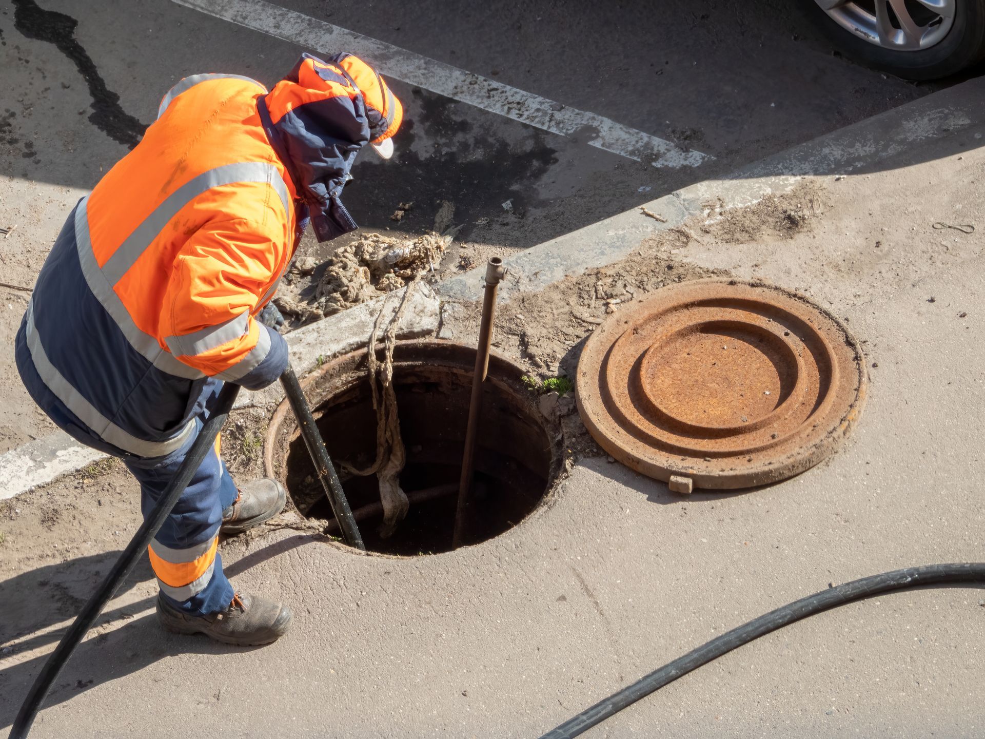 Worker in orange safety gear inspecting an open manhole on a street.