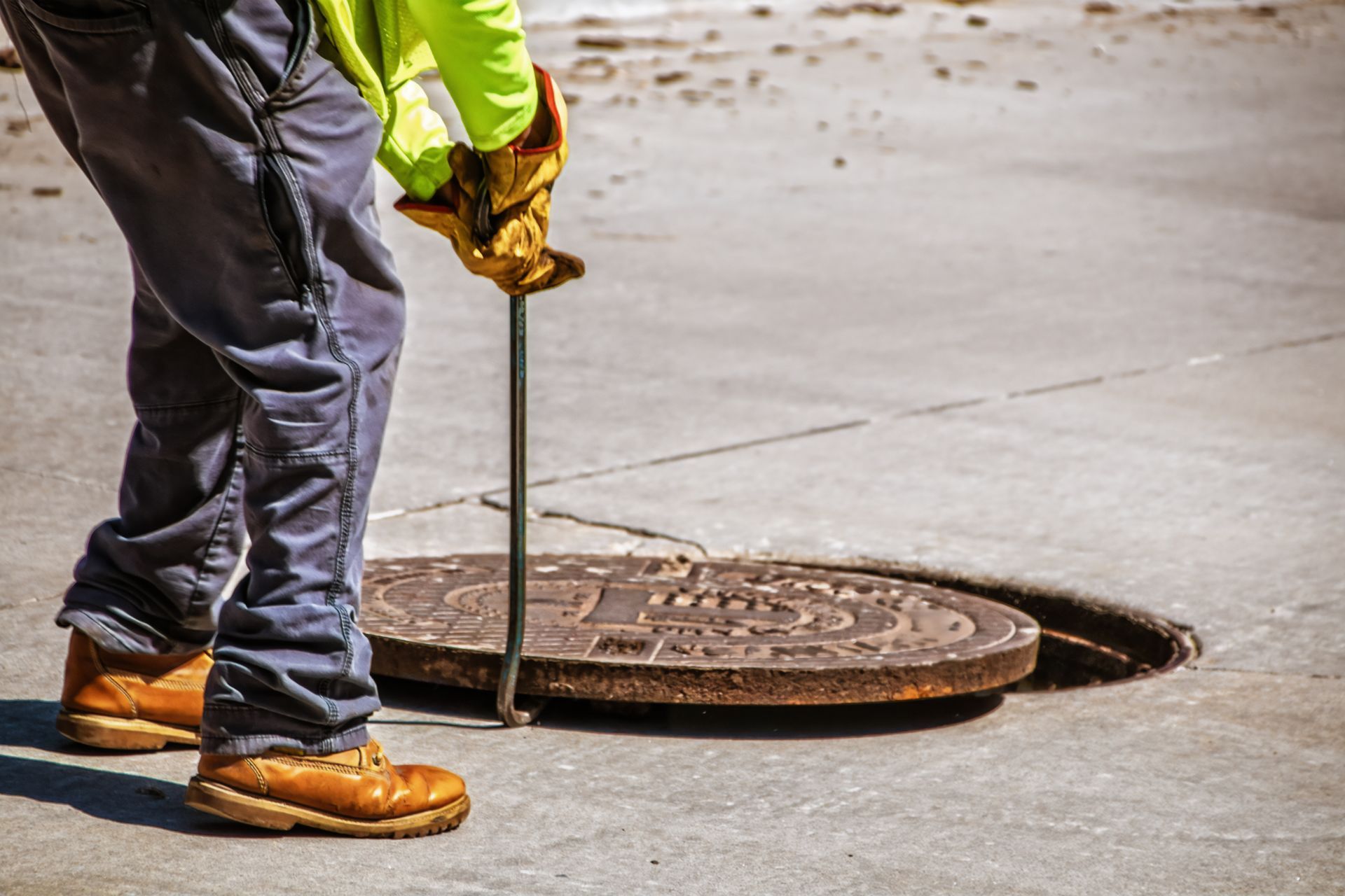 Worker in reflective jacket lifting a manhole cover with a pry bar on a concrete street