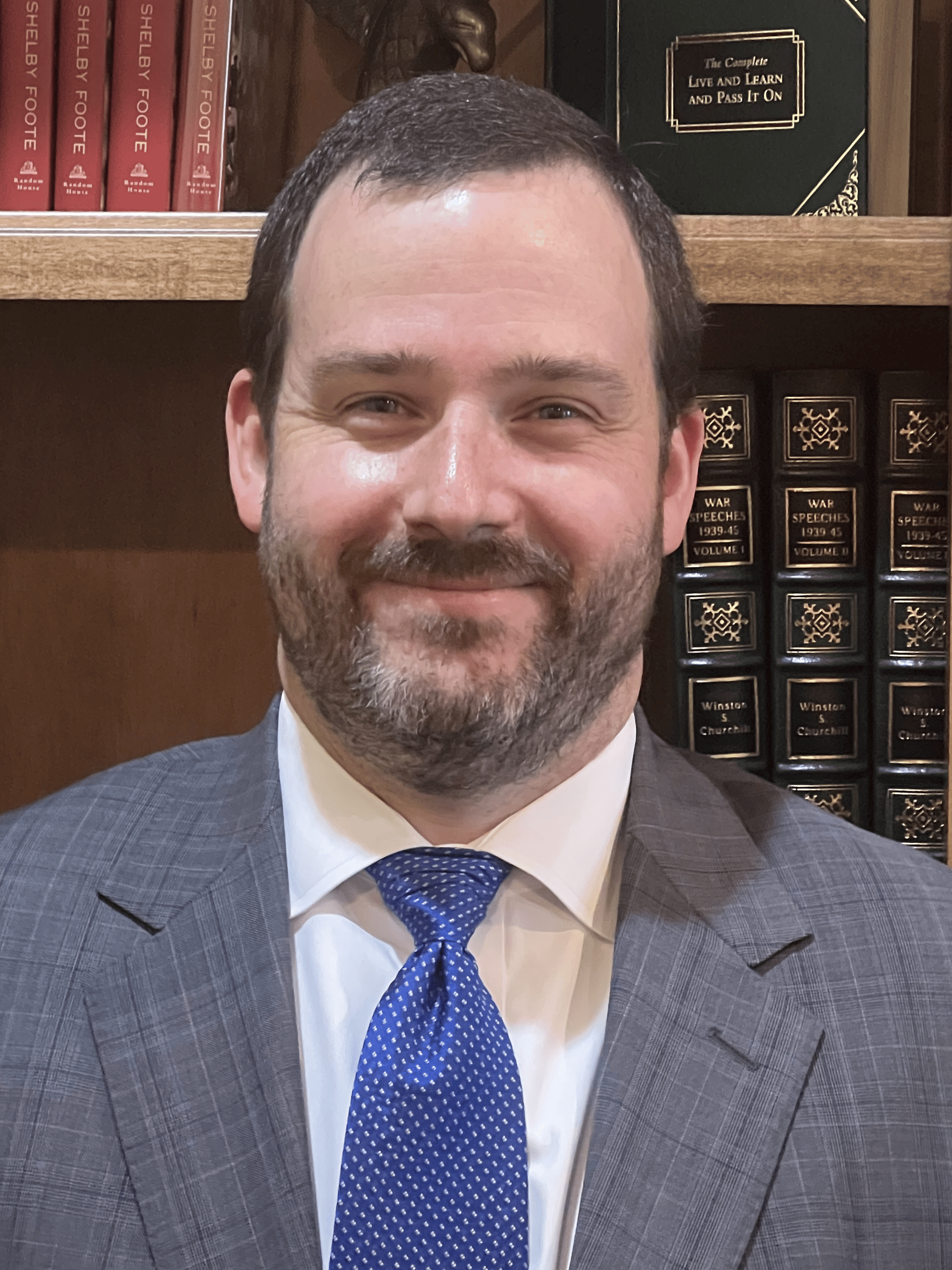 A man in a suit and tie is standing in front of a bookshelf filled with books.