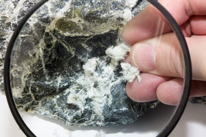 Hand holding a magnifying glass over a dark rock with white crystalline veins and flecks