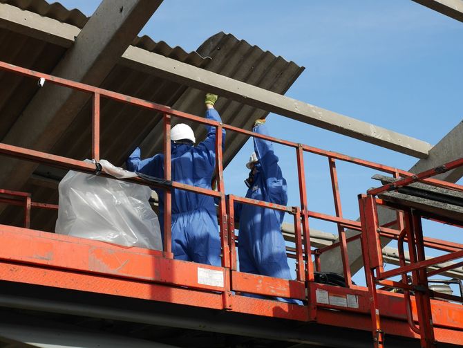 Workers in blue coveralls and hard hats on a red lift, installing roof panels on a building