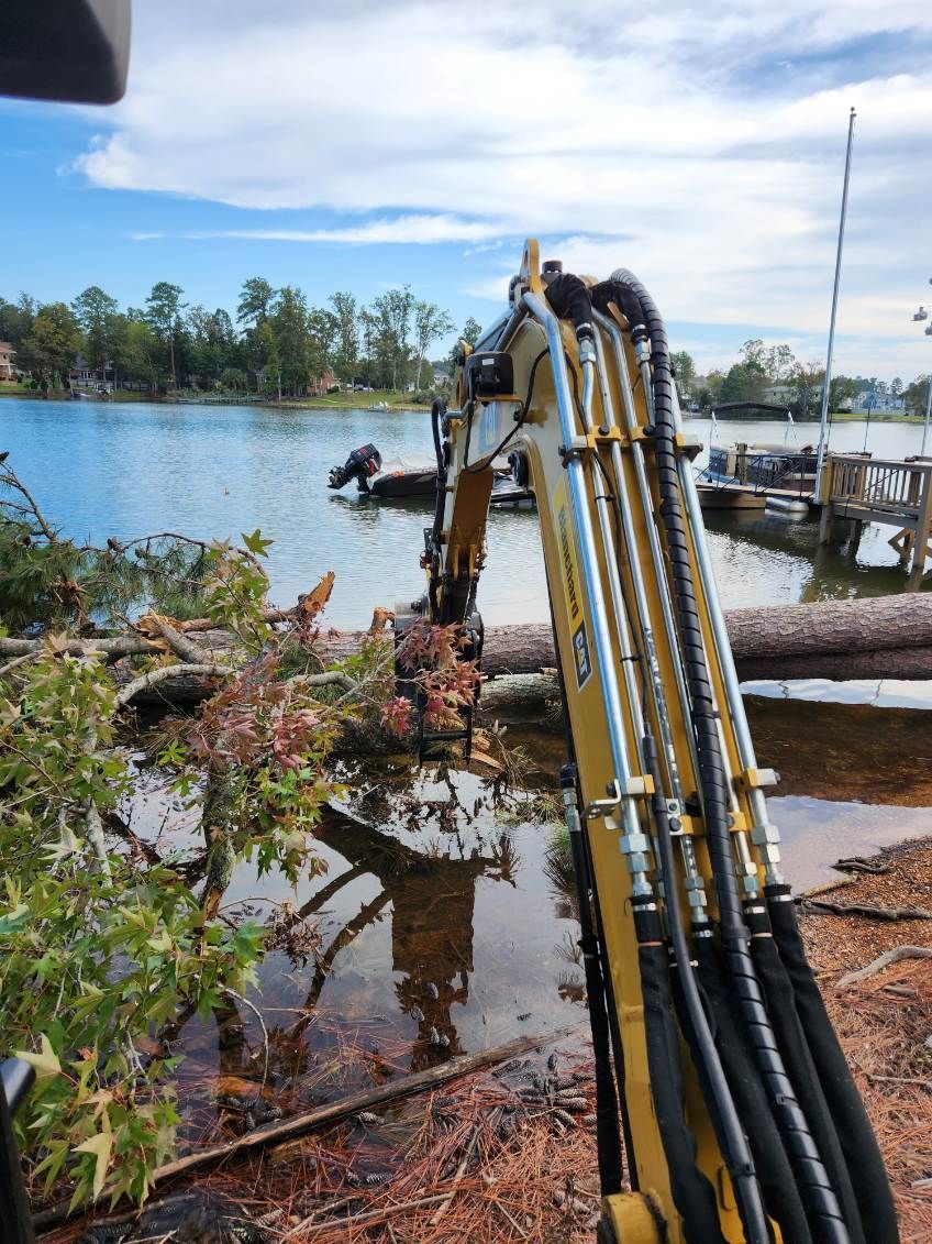 An excavator removing a fallen tree from a lake. The sky is blue with fluffy clouds.