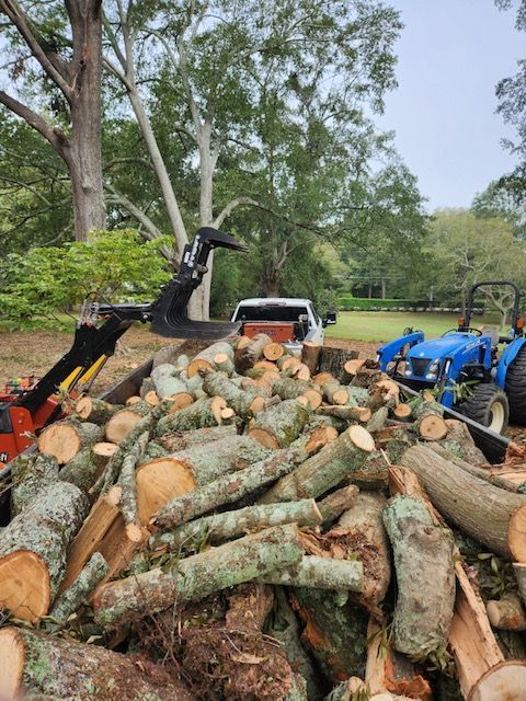 Logs piled near a blue tractor and truck; a mechanical claw holds more logs in a wooded area.