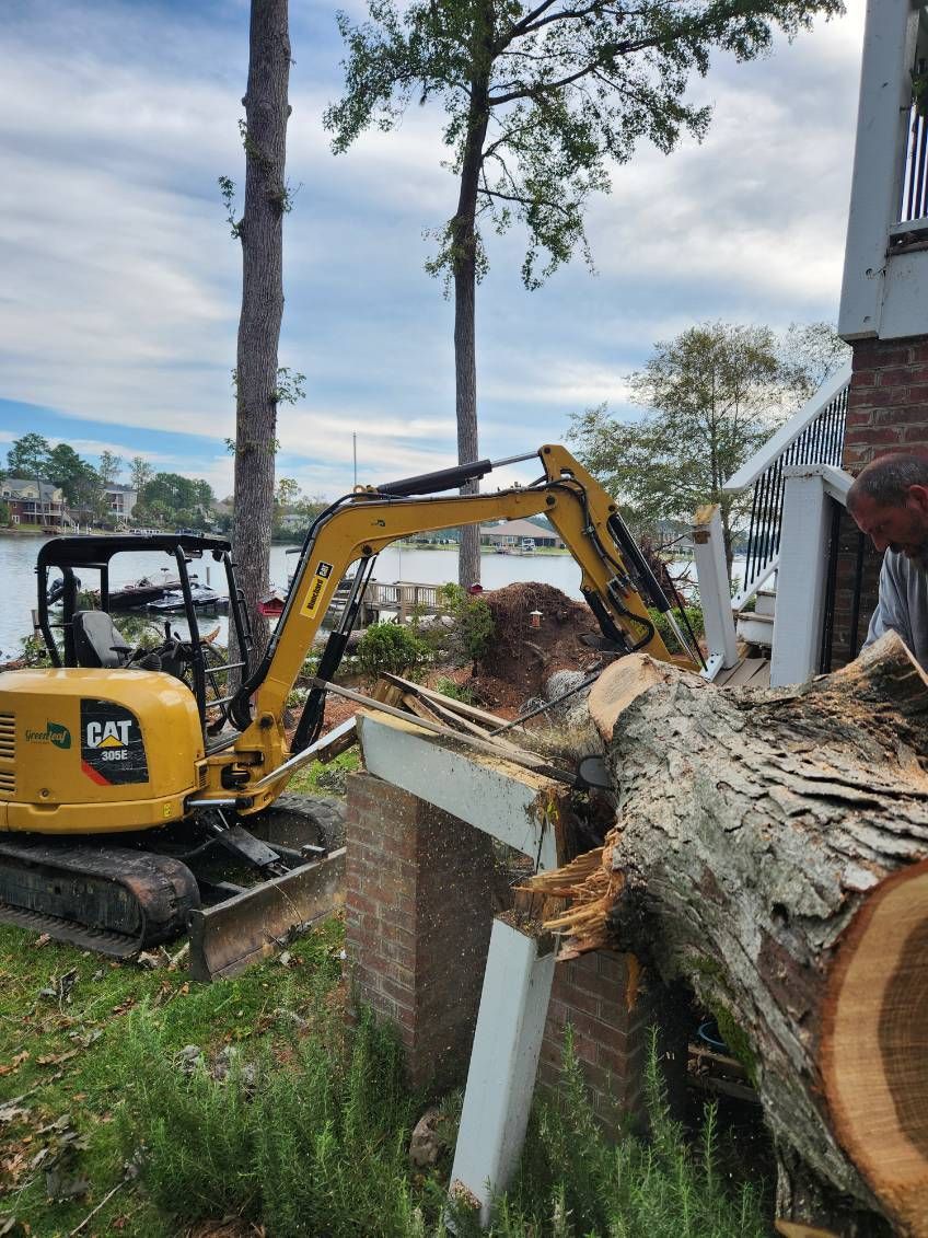 Yellow excavator removing a tree branch near a brick structure. Lake and trees in the background.