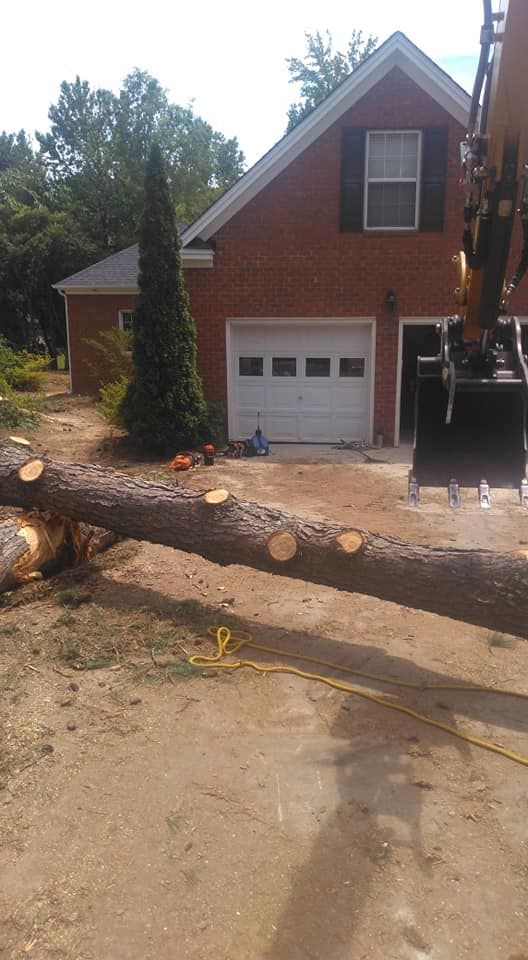 A fallen tree trunk in front of a brick house with a garage; an excavator is visible.