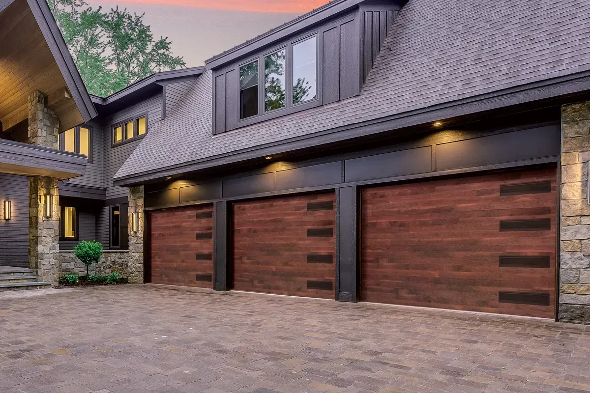 A house exterior with three wood-look garage doors, dark trim, stone pillars, and a dormer window under a sunset sky.