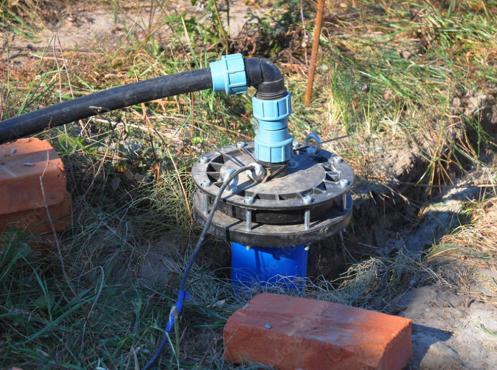 Photograph of a water pump installation outdoors, showing a black hose connected to a blue and black