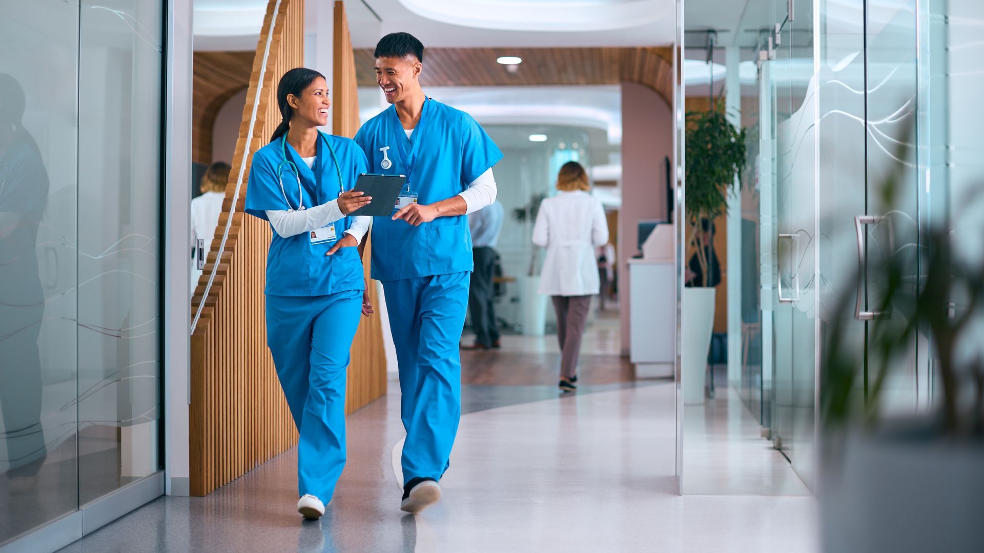 A man and a woman are walking down a hospital hallway looking at a tablet.
