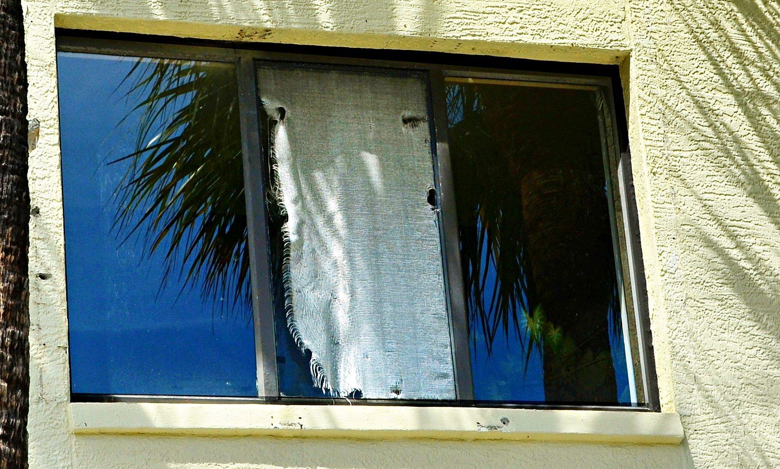 Damaged window with a broken pane and torn curtain on a beige stucco wall