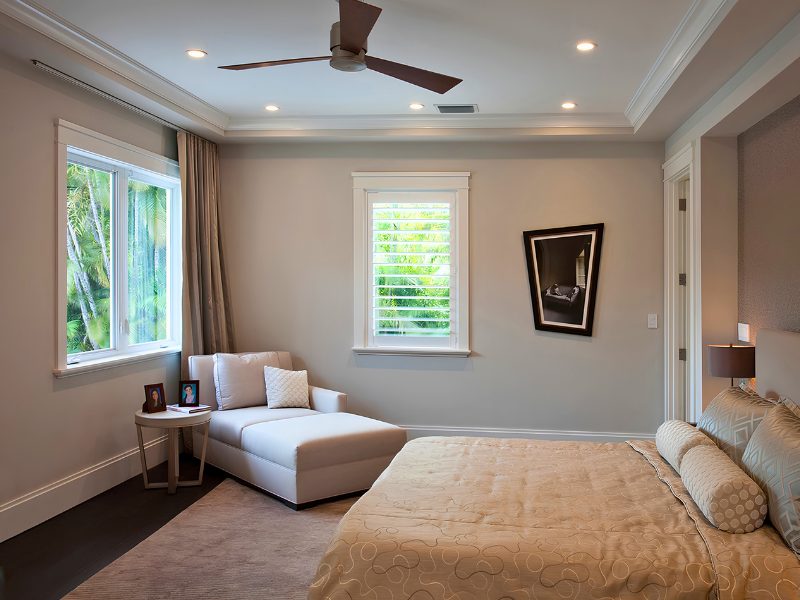 Bright modern bedroom with beige walls, a large bed, chaise lounge, and two windows with natural light.